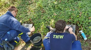 Auf dem Foto sitzen zwei Jugendliche vor zwei Kriegsgräbern. Mit einem Pflegemittel sowie einer Bürste schrubben sie die Grabplatten im Rasen. Von Ihnen sind lediglich die Rücken und bei einem Jungen die große gelbe Aufschrift „THW“ auf der dunklen T-Shirt-Rückseite zu erkennen.