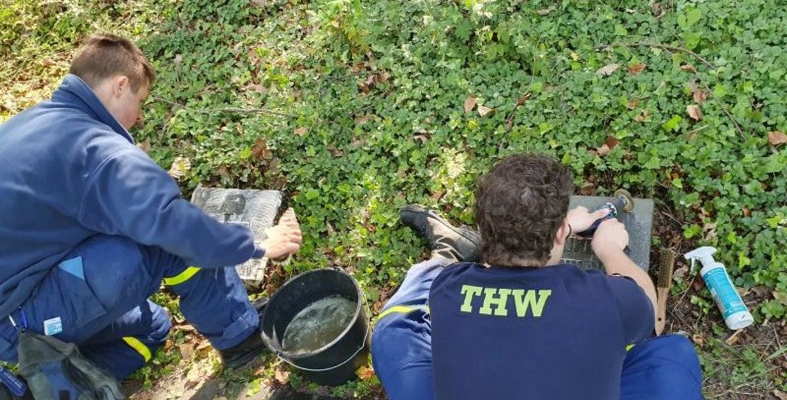 Auf dem Foto sitzen zwei Jugendliche vor zwei Kriegsgräbern. Mit einem Pflegemittel sowie einer Bürste schrubben sie die Grabplatten im Rasen. Von Ihnen sind lediglich die Rücken und bei einem Jungen die große gelbe Aufschrift „THW“ auf der dunklen T-Shirt-Rückseite zu erkennen.